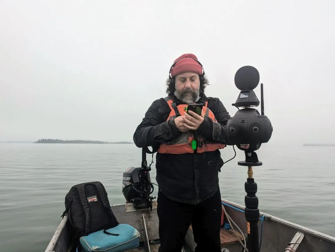 Pablo Saravanja in a red toque and life jacket, checking his phone on a small boat in the fog, beside a 360-degree camera rig on a pole.